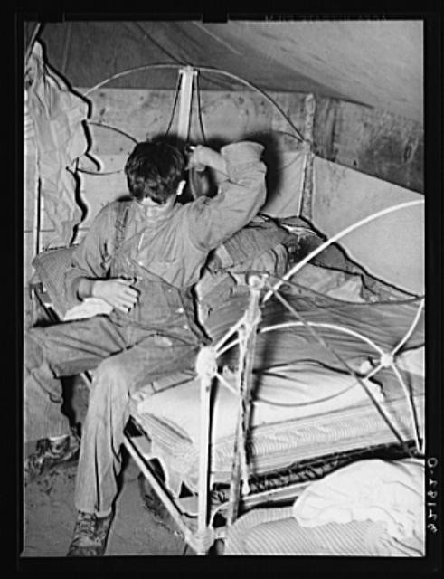 Son of white migrant worker putting on overalls in tent...