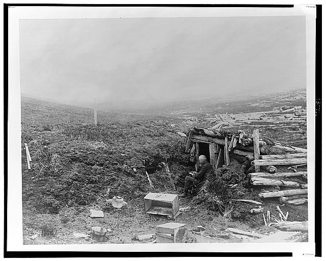 [Unidentified old man, seated, writing, at entrance to a...