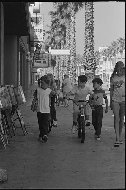 [Children walking on sidewalk, by news racks selling...