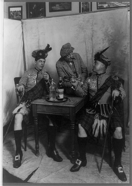 [Two men wearing kilts seated at small table, drinking...