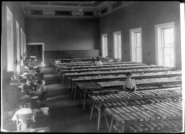 [Long tables piled with cards and people working in the...