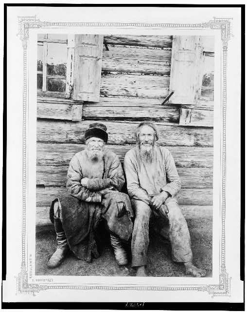 [Two men seated, outside of log house, full-length,...
